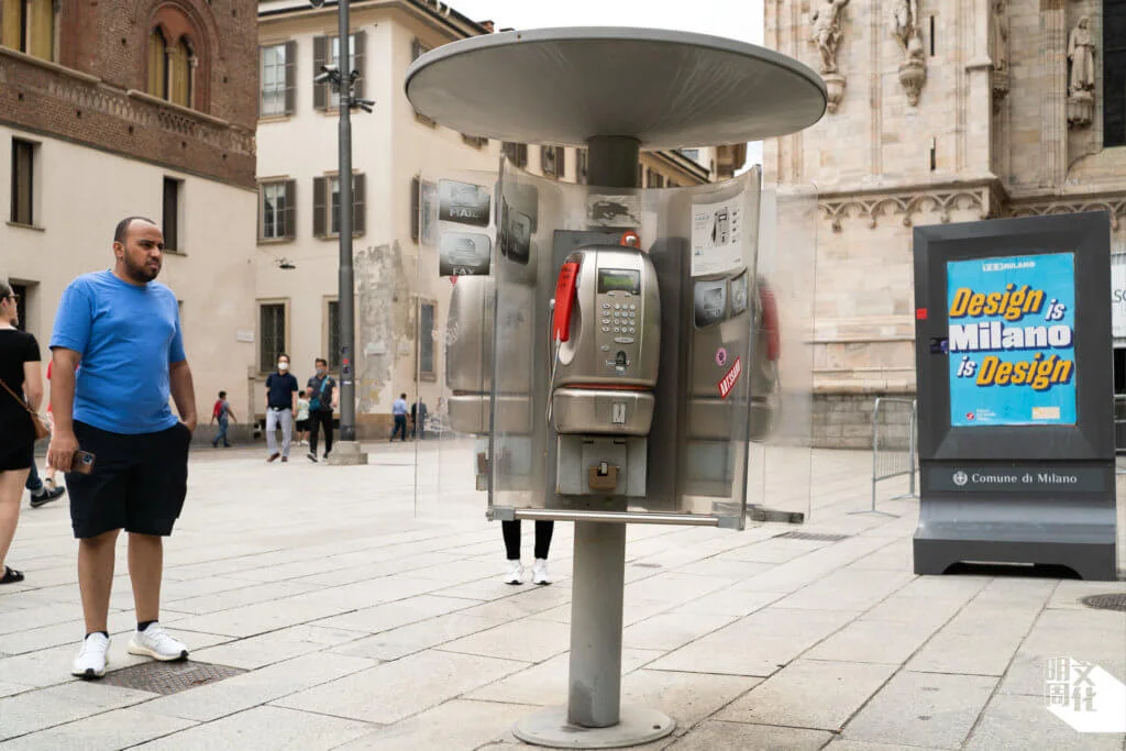 米蘭王宮（Palazzo Reale di Milano）旁邊的舊式電話亭，左側男子一面錯愕，右側廣告牌則在宣傳米蘭設計周。