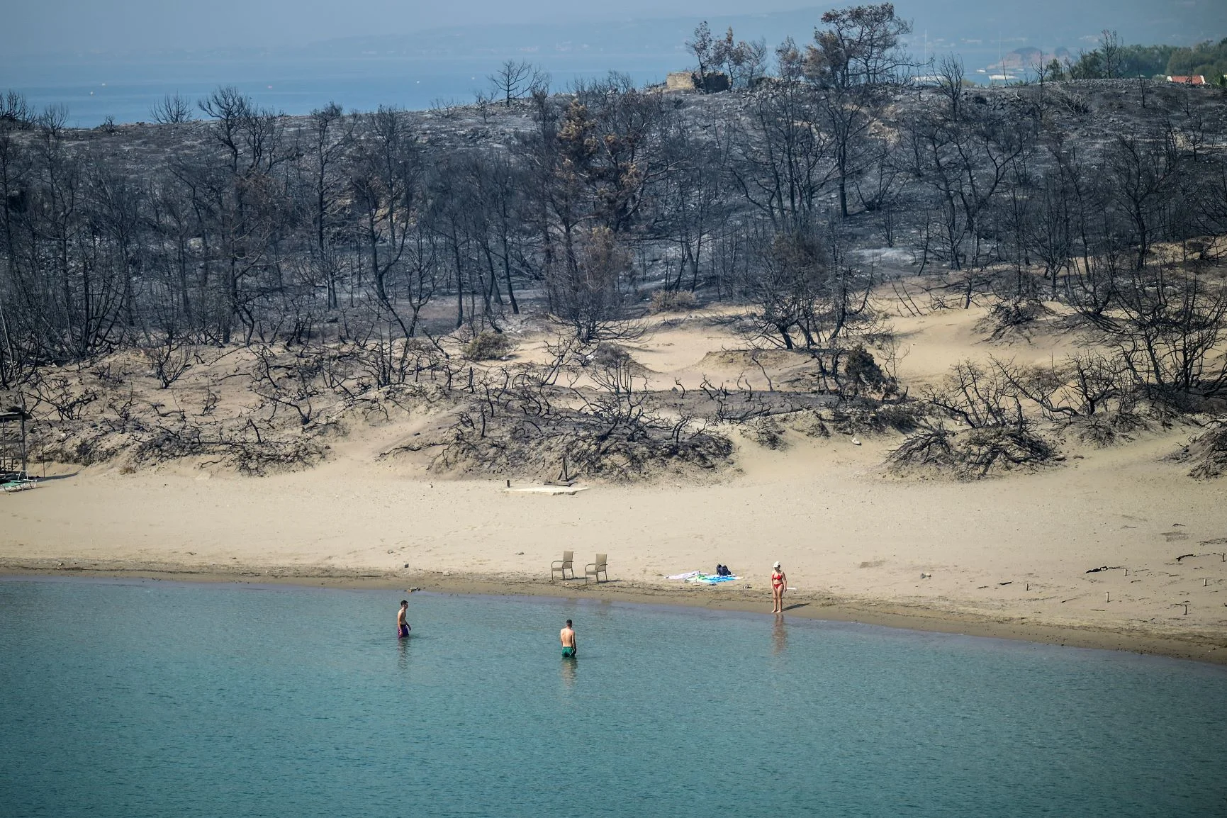 酷熱天氣導致希臘野火肆虐，當地七月曾發生嚴重山火。圖中女子身處 希臘羅得島（Rhodes）一個沙灘，她身後是一片遭野火燒毀的樹林。（ANGELOS TZORTZINIS / AFP）