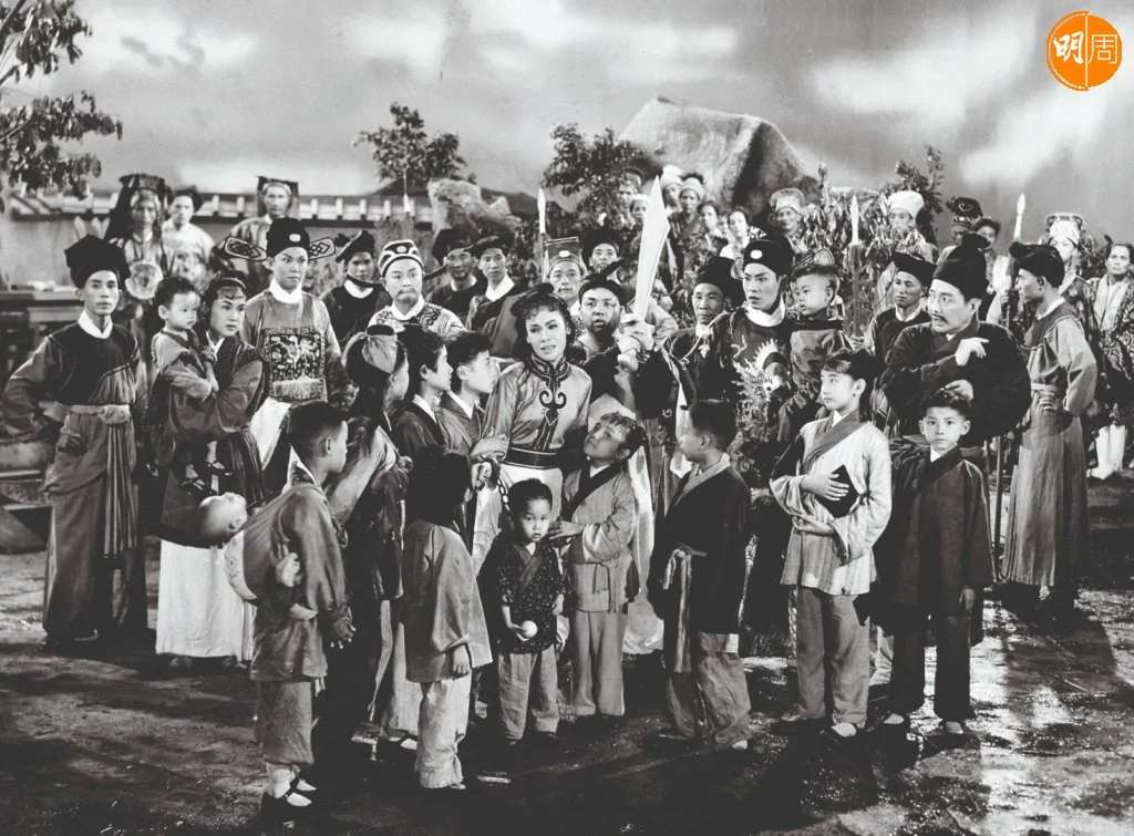 《七兒八女九狀詞》（1960）的一幀劇照裏，女主角余麗珍身披鐐鎖被數不清楚到底多少的童星圍住。她和男主角羅劍郎的表情皆很入戲，照片高度還原現場的分毫，左上角的衙差、居中及右下角的童星，不約而同望向鏡頭與觀眾對上眼。（圖片提供：任國光）